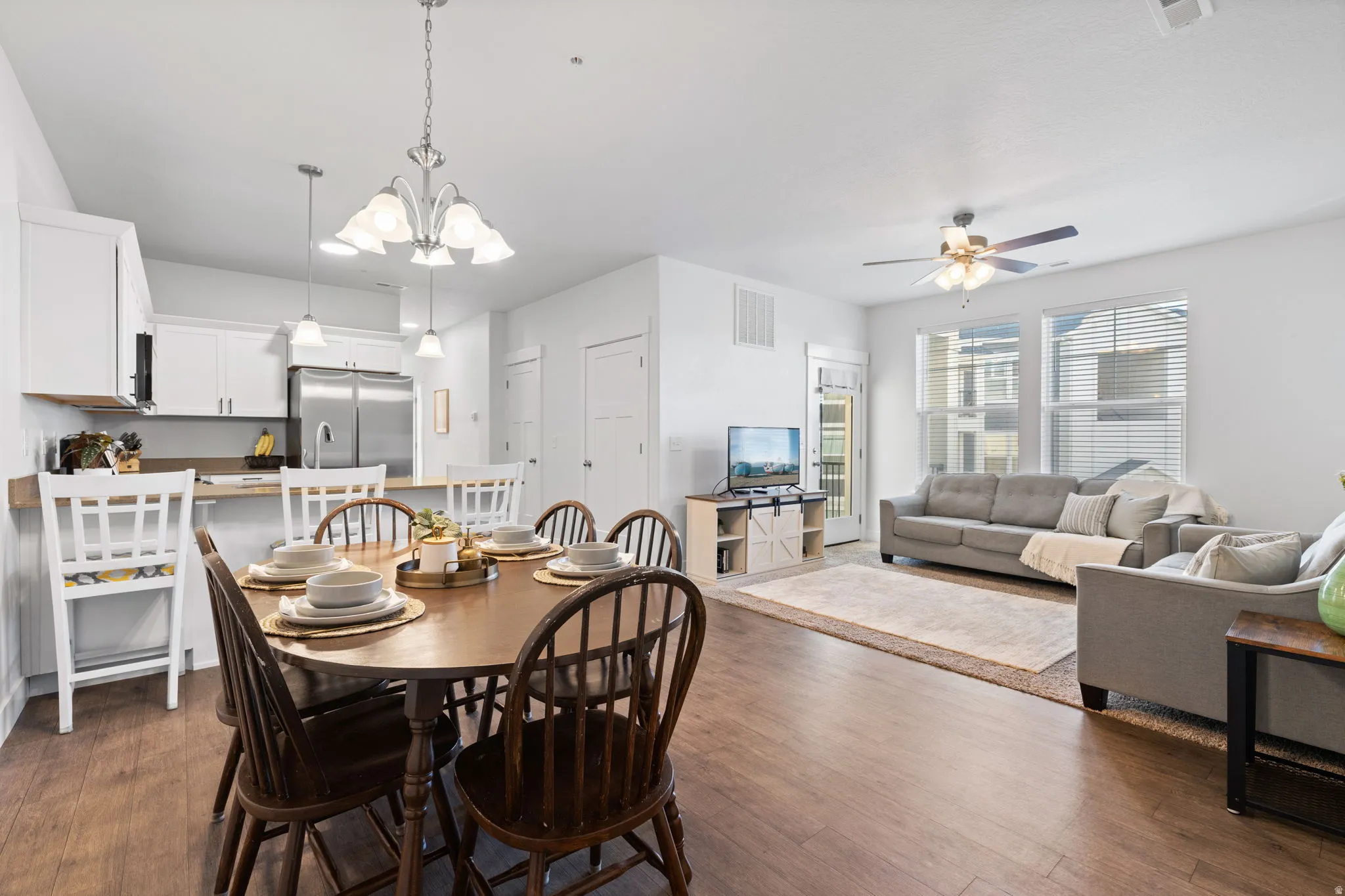 Open-concept living space featuring wood-finish flooring, a contemporary kitchen with white cabinetry and stainless steel appliances, and a dining area illuminated by a decorative chandelier