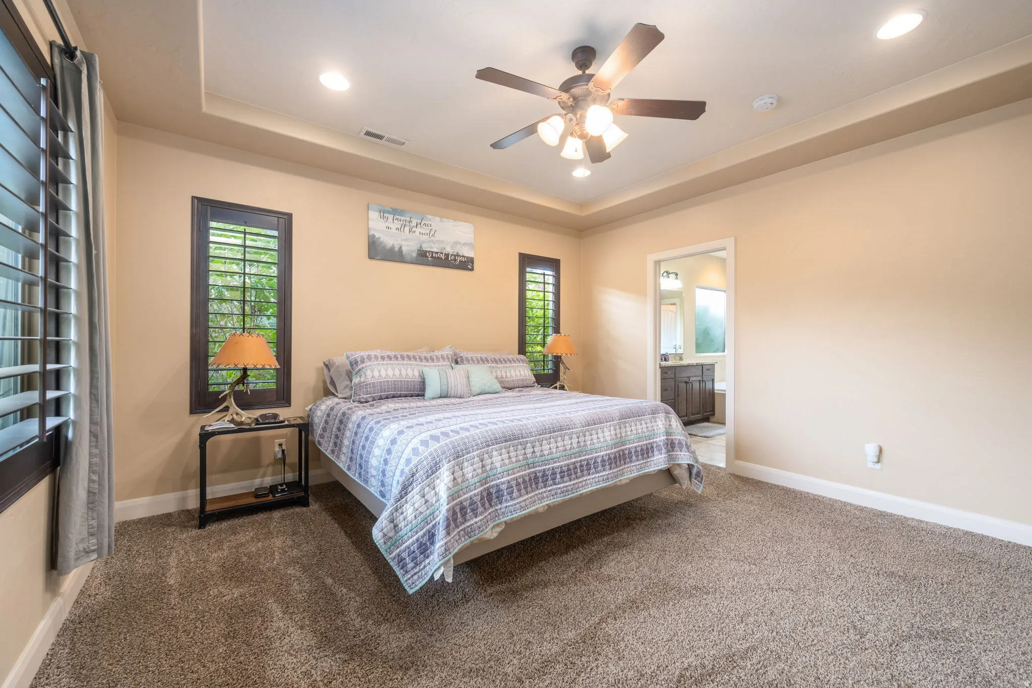 Carpeted bedroom with a raised ceiling, recessed lighting, ceiling fan, and ensuite bath