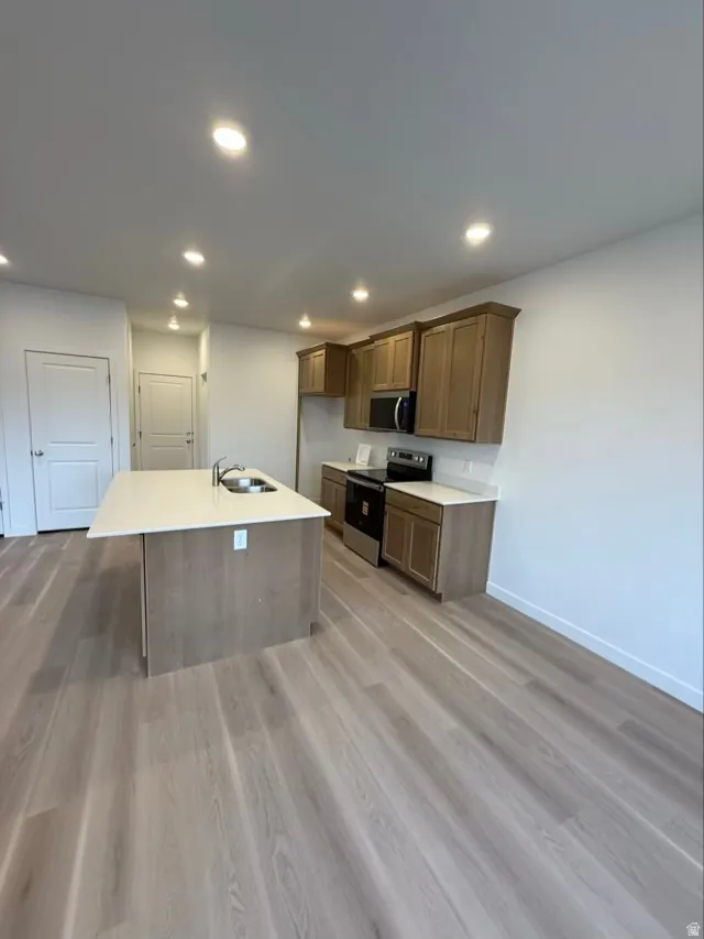 Kitchen featuring stainless steel appliances, light wood finished floors, a kitchen island with sink, recessed lighting, and brown cabinets