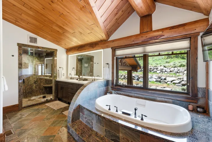 Spacious bathroom featuring a built-in soaking tub, a large window, a glass-enclosed shower, a double vanity with an illuminated mirror, and exposed wood beams