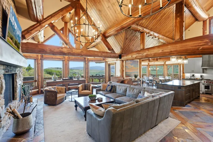 Vaulted wood-paneled ceilings with exposed log beams, floor-to-ceiling windows, stone-faced fireplace, large kitchen island with dark countertop, and integrated stainless steel appliances