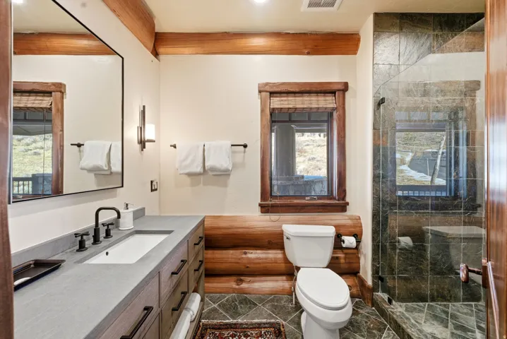 Bathroom featuring a wood-finish vanity with an integrated sink and matte black fixtures, a large mirror, a glass-enclosed shower with stone-finish tile, and exposed wood beam accents