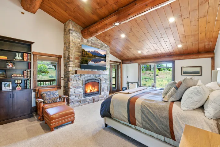 Vaulted wood-plank ceiling with exposed log beams, stone-faced fireplace with wood mantel, built-in shelving with lower cabinetry, light-colored carpeting, and large windows