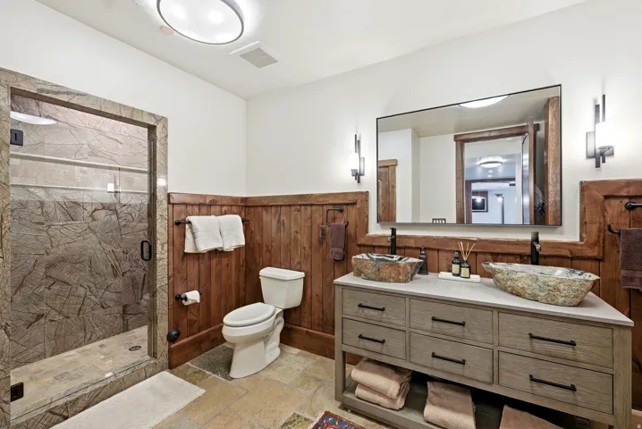 Bathroom featuring a glass-enclosed shower with stone surround, rustic wood paneling, a dual vanity with vessel sinks, and stone tile flooring