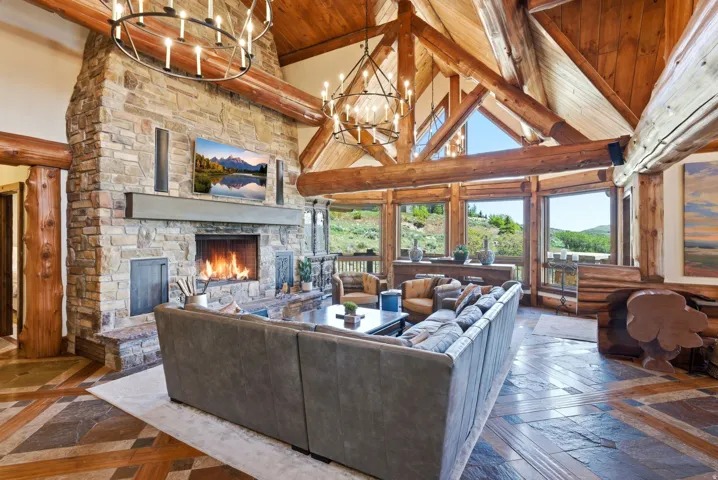 Grand living space featuring a stone fireplace with a substantial hearth, vaulted wood-beam ceilings, and extensive window walls capturing the natural landscape