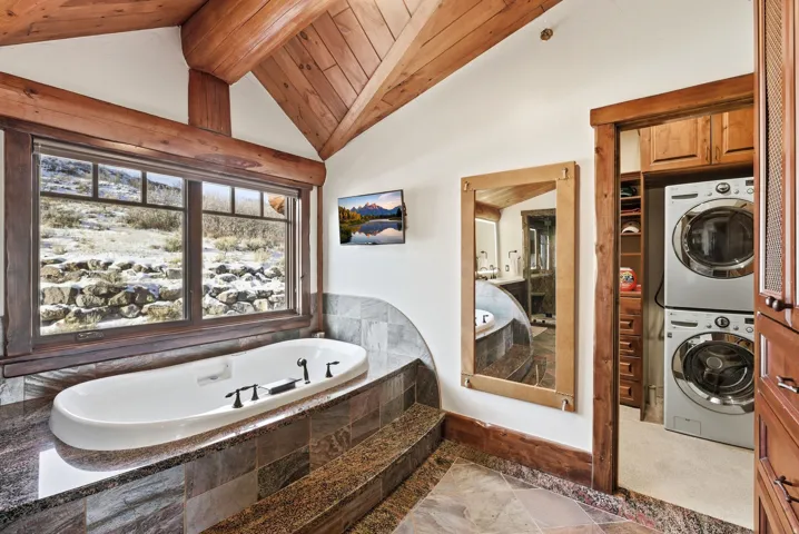 Freestanding soaking tub with dark fixtures set on a stone-clad platform, under a large window with natural light