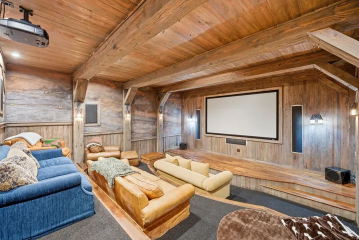 Dedicated home theater featuring a large projection screen, integrated projector, wood-finish wall paneling, exposed wood beams, and tiered flooring