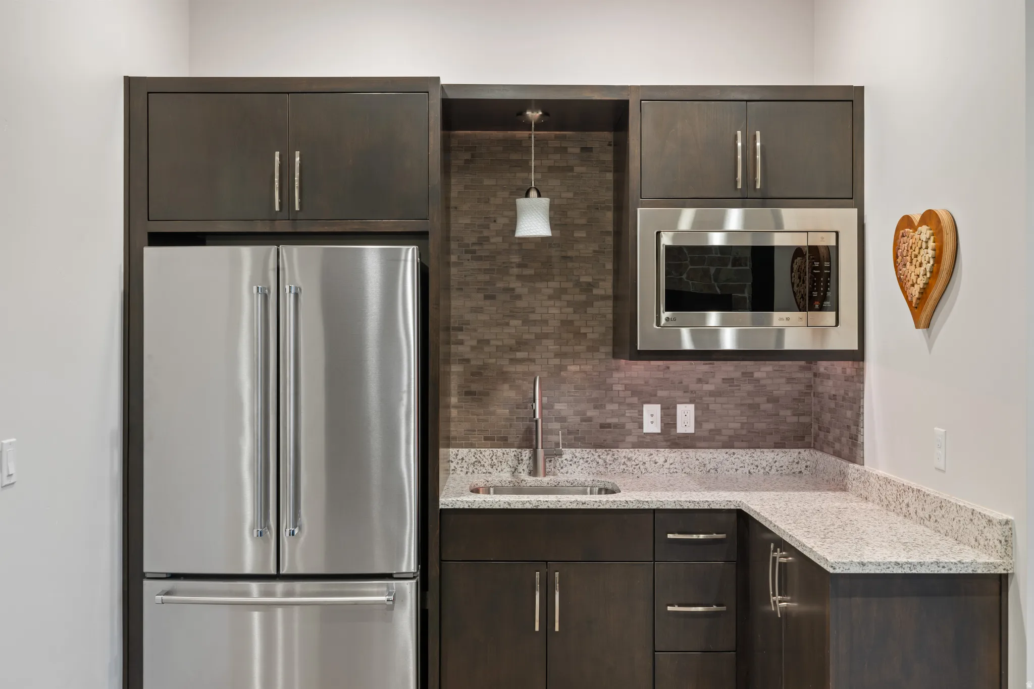 Kitchen featuring dark brown cabinetry, stainless steel appliances, light stone countertops, tasteful backsplash, and decorative light fixtures