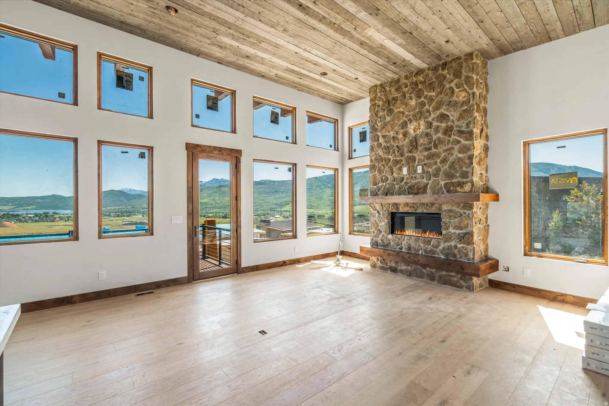 Spacious room featuring a floor-to-ceiling stone fireplace with a wood mantle and electric insert, multiple large windows with wood trim, and a wood-finish ceiling
