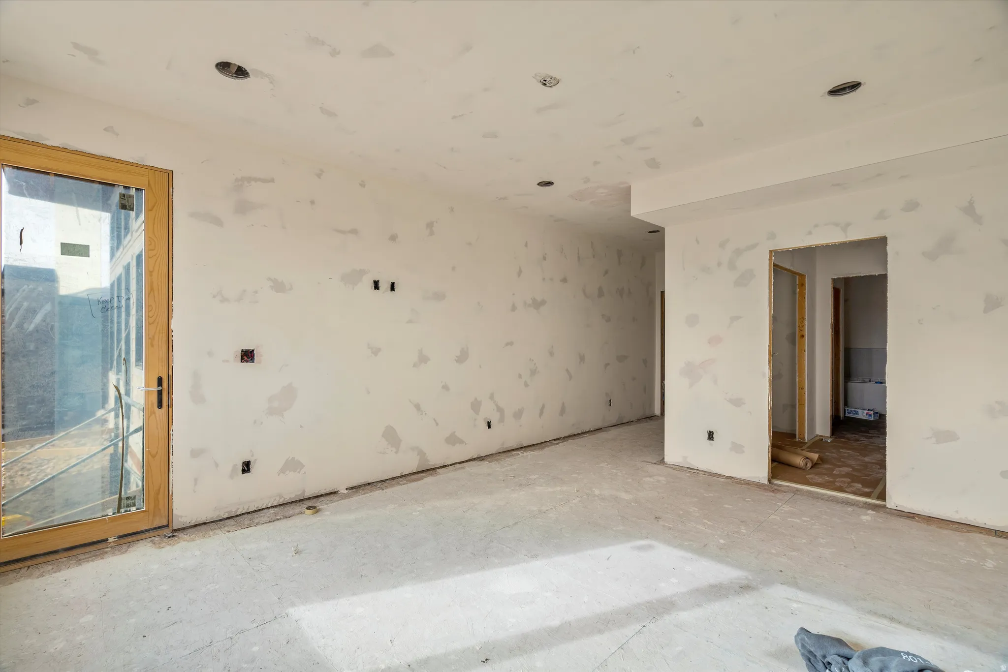 Interior space showcasing a large glass door with a light wood frame, multiple recessed light fixtures, and raw drywall finishes