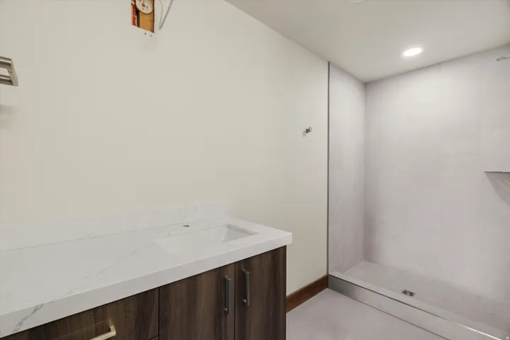 Modern primary bathroom featuring a wood-finish vanity with dual cabinet storage, white quartz countertop, and frameless glass shower enclosure with white subway tile surround and linear floor drain