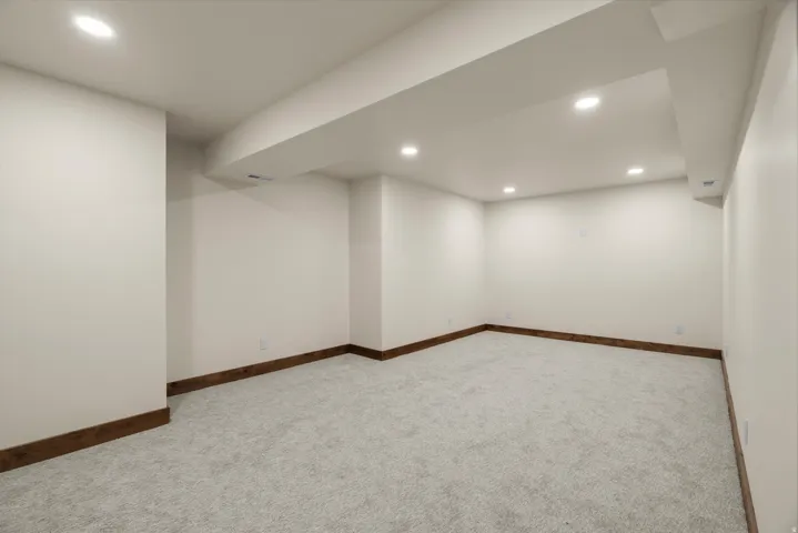 Spacious basement room featuring recessed ceiling lighting, smooth drywall finishes, and neutral carpet flooring with wood-trim baseboards throughout