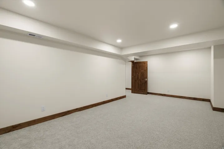 Spacious finished room featuring recessed ceiling lighting, clean white walls, neutral carpet flooring, and rich wood-finish trim accents