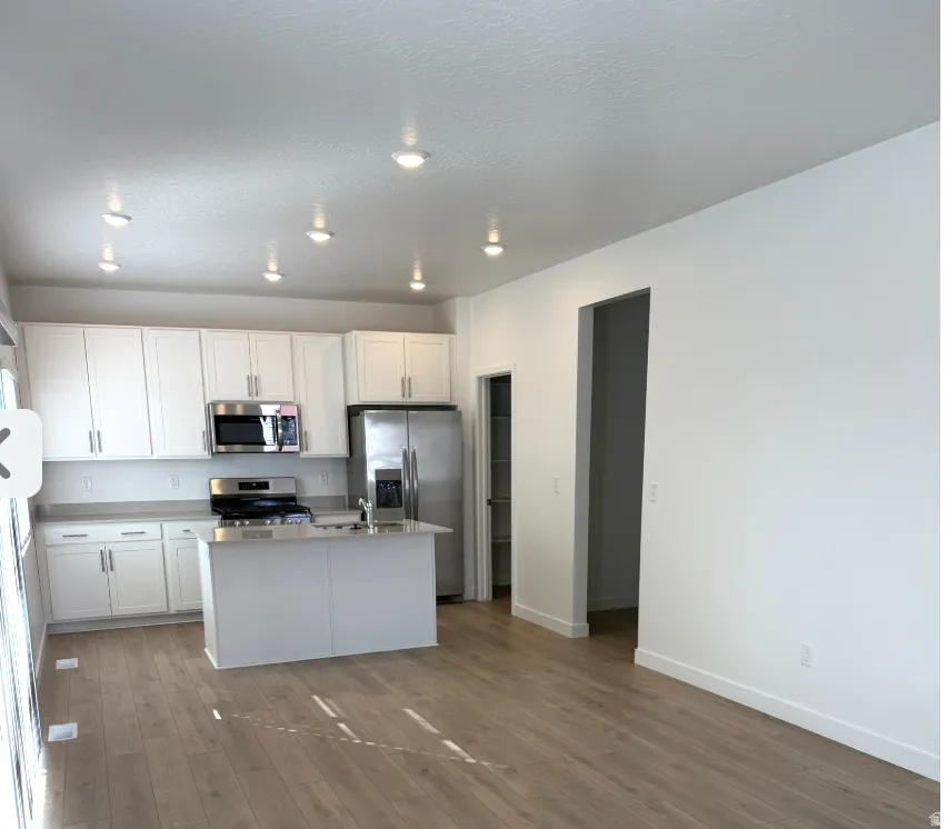 Kitchen featuring white cabinets, appliances with stainless steel finishes, a center island with sink, light wood finished floors, and light stone counters