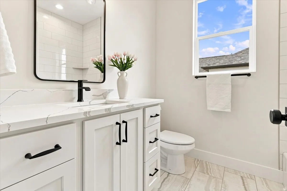 Modern bathroom featuring a white vanity with dual storage drawers and black hardware, a rectangular framed mirror, quartz countertop, matte black wall-mounted faucet, porcelain toilet, wood-finish flooring, and natural light from a transom window