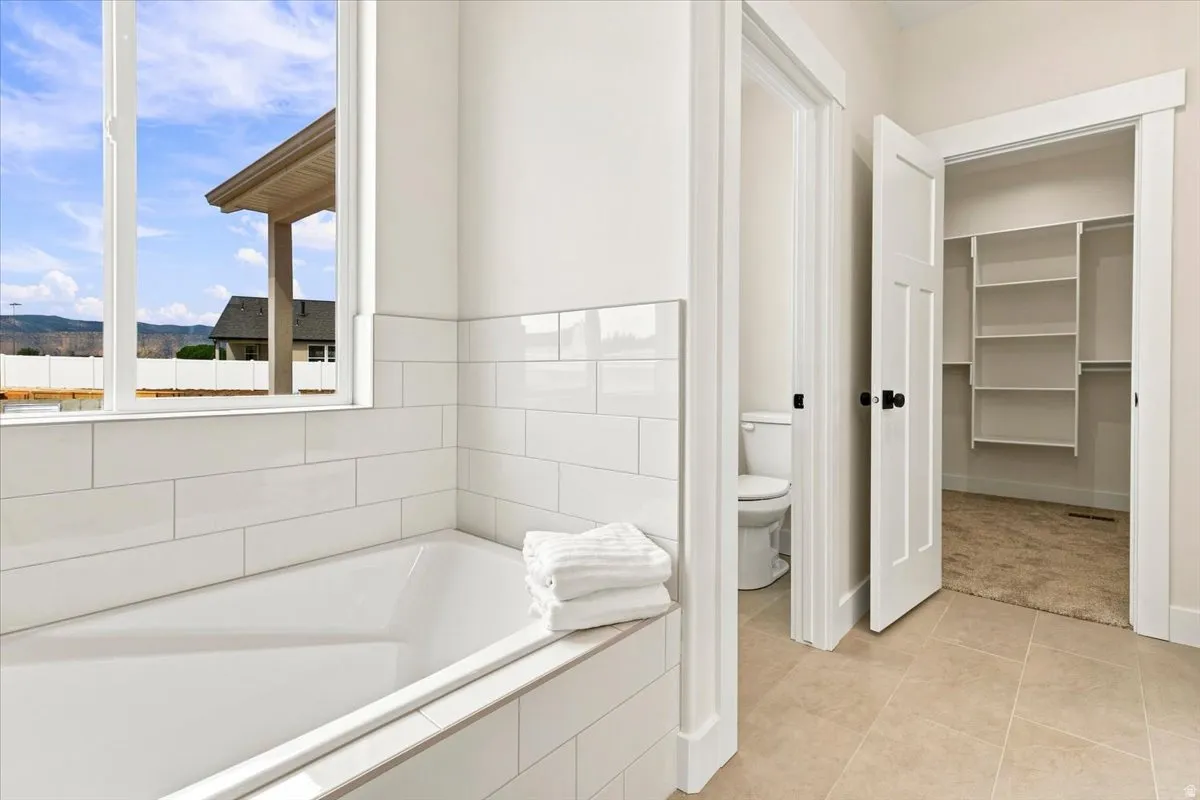 Freestanding bathtub with subway tile surround, large window, tile flooring, separate water closet, and walk-in closet with built-in shelving