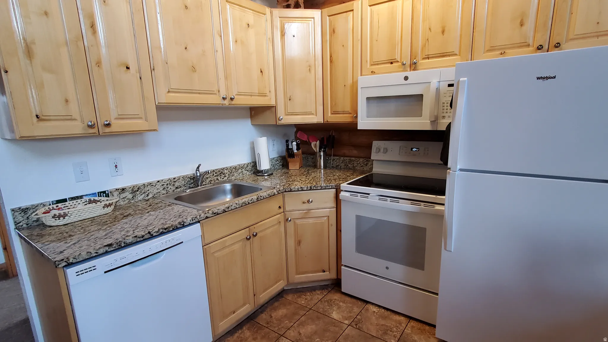 Kitchen featuring white appliances, light brown cabinets, dark tile patterned flooring, and dark stone counters