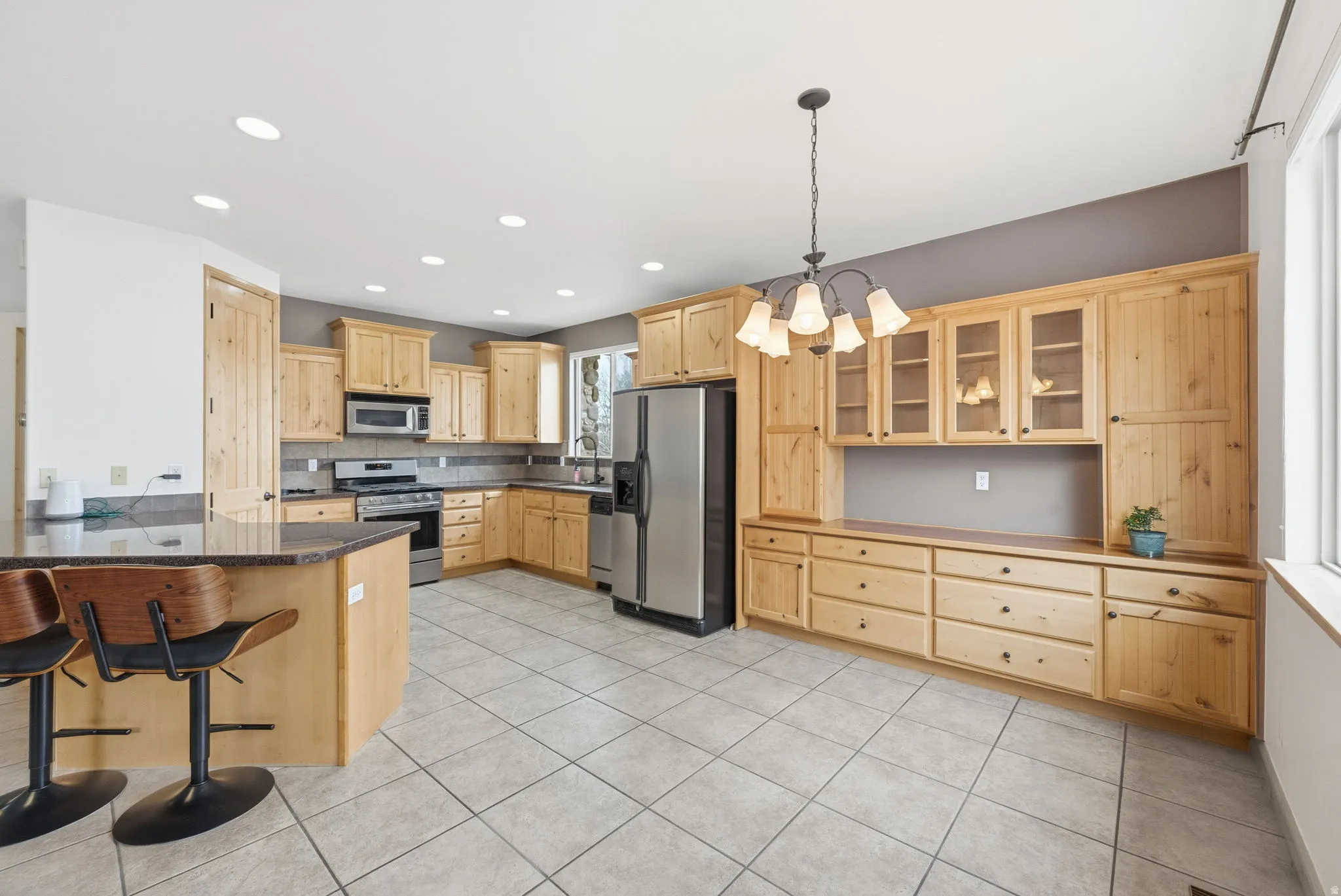 Kitchen with light brown cabinetry, glass insert cabinets, appliances with stainless steel finishes, a breakfast bar area, and a chandelier