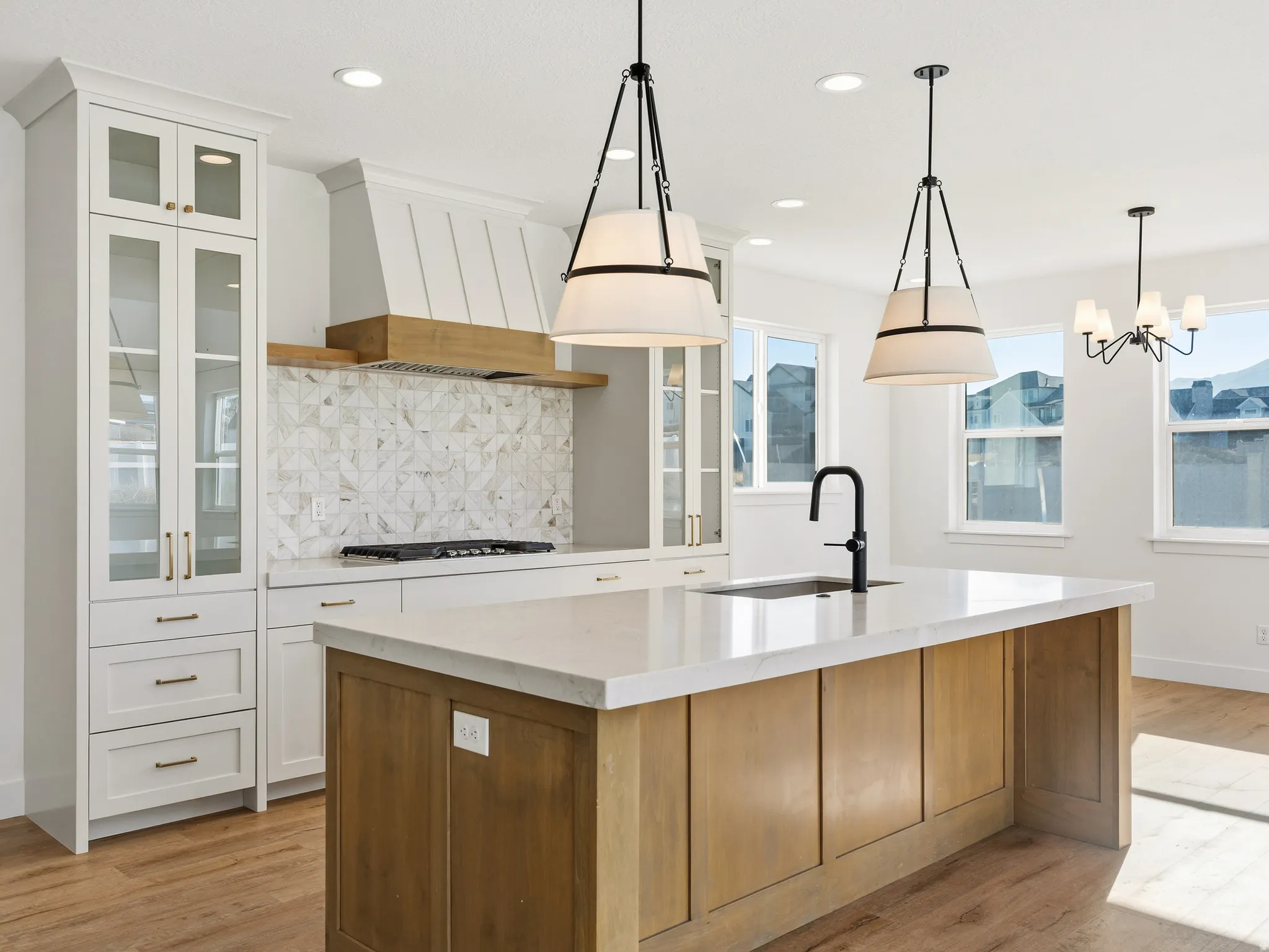 Two tone kitchen with two tone cabinetry, light stone countertops, glass insert cabinets, tasteful backsplash, and a center island with sink