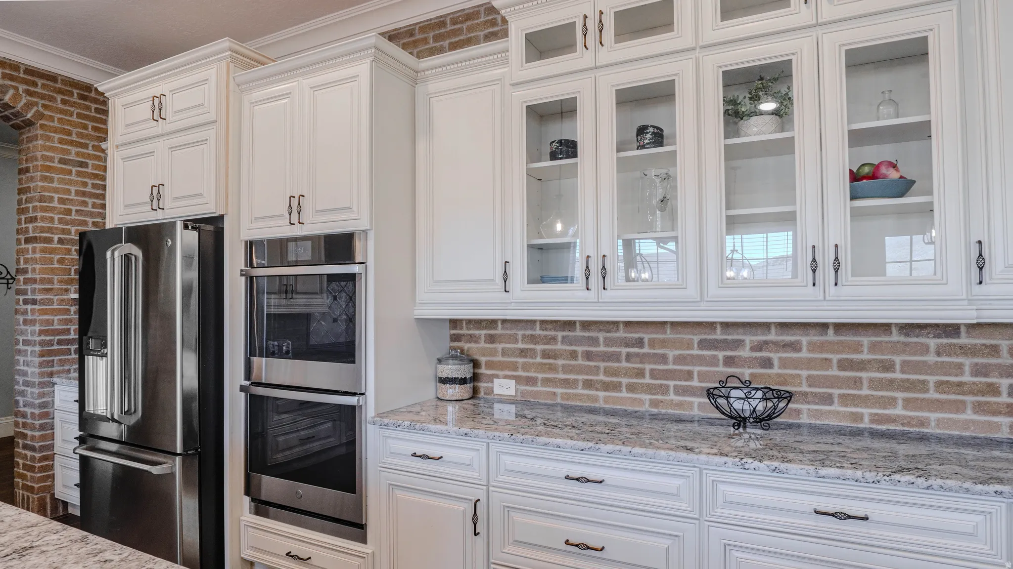 Kitchen featuring stainless steel appliances, light stone countertops, white cabinets, glass insert cabinets, and brick wall