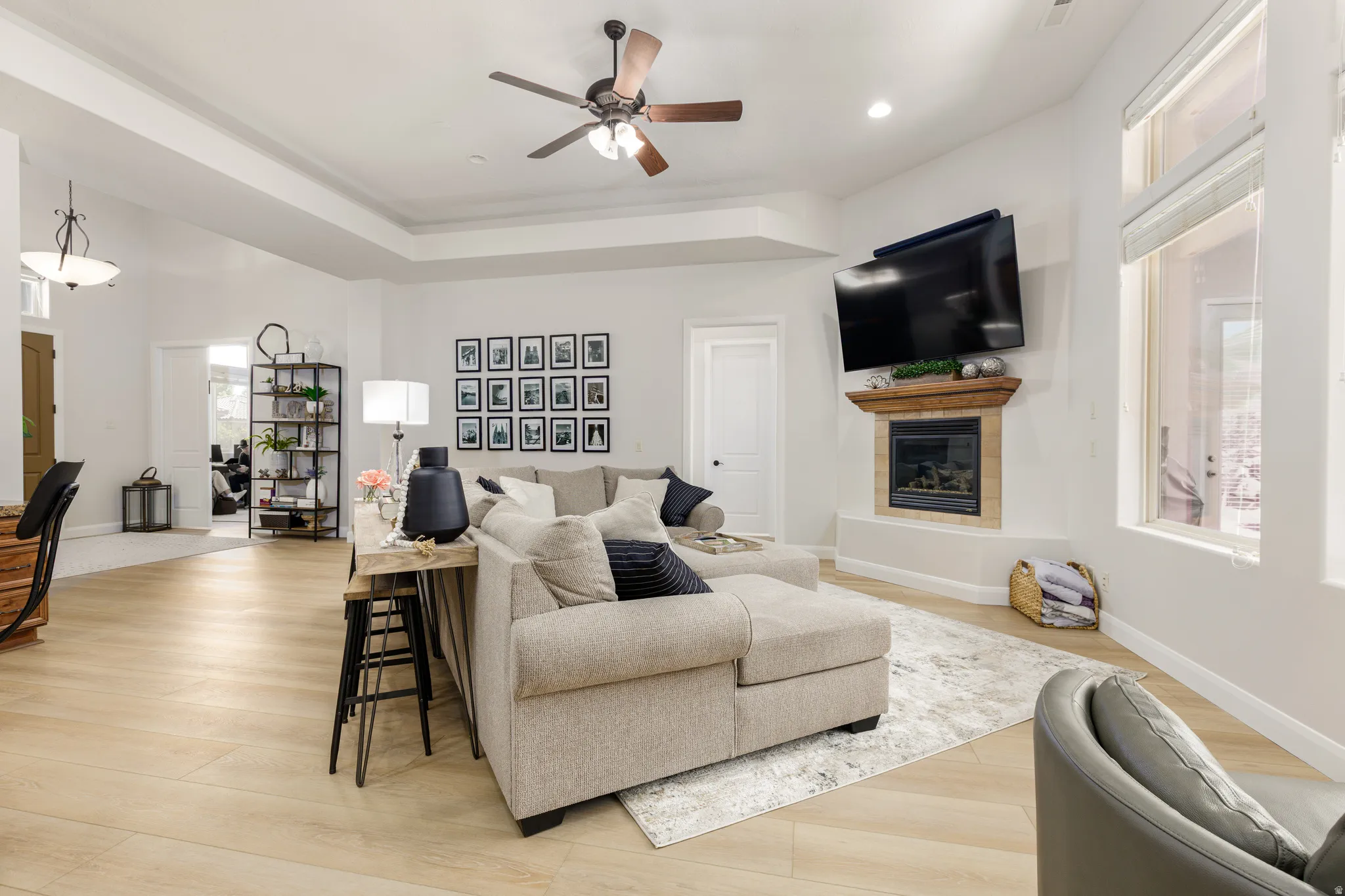 Living area with ceiling fan, a tiled fireplace, light wood-type flooring, and a raised ceiling