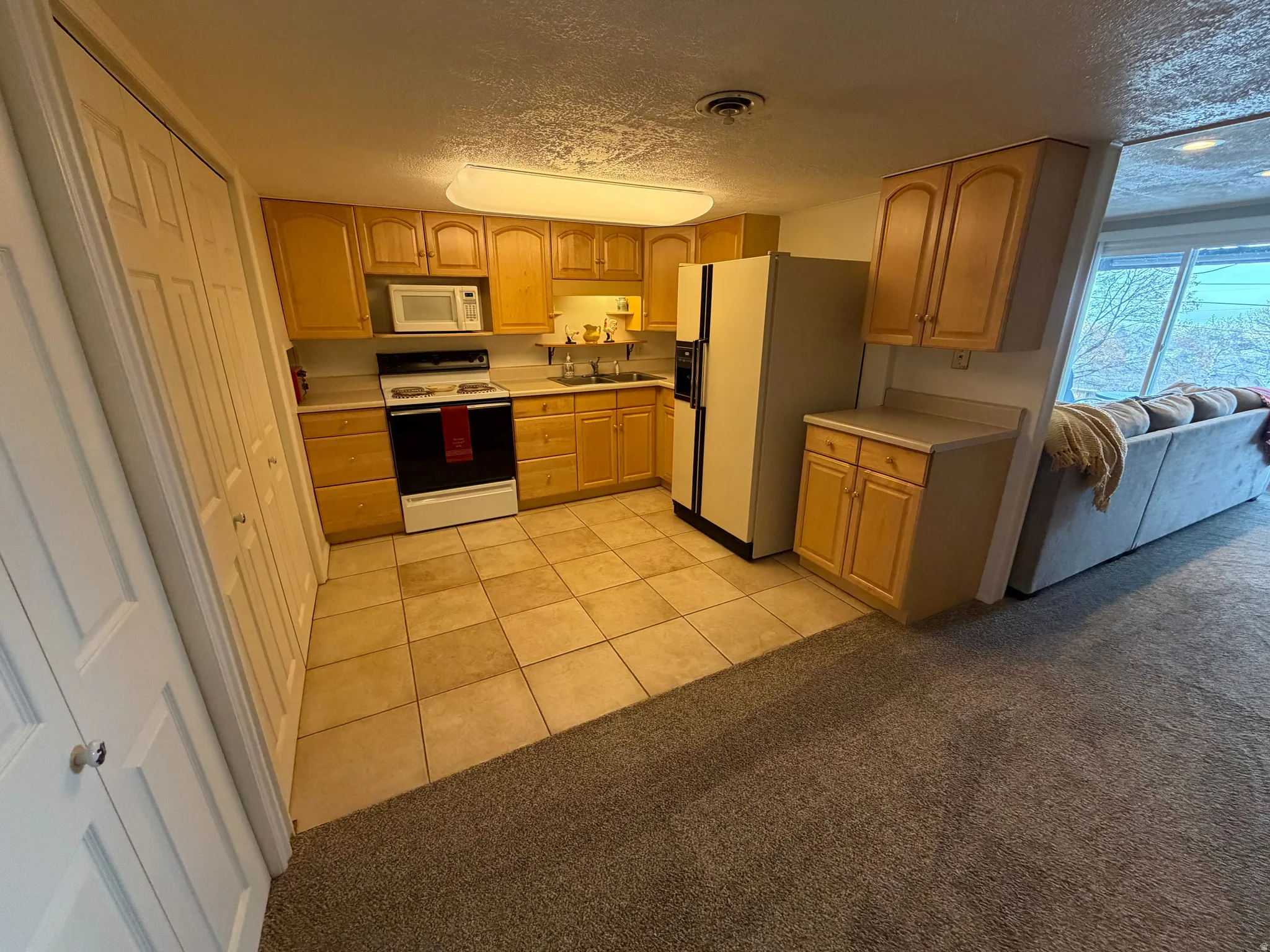 Kitchen with light countertops, a textured ceiling, white appliances, light carpet, and light brown cabinetry
