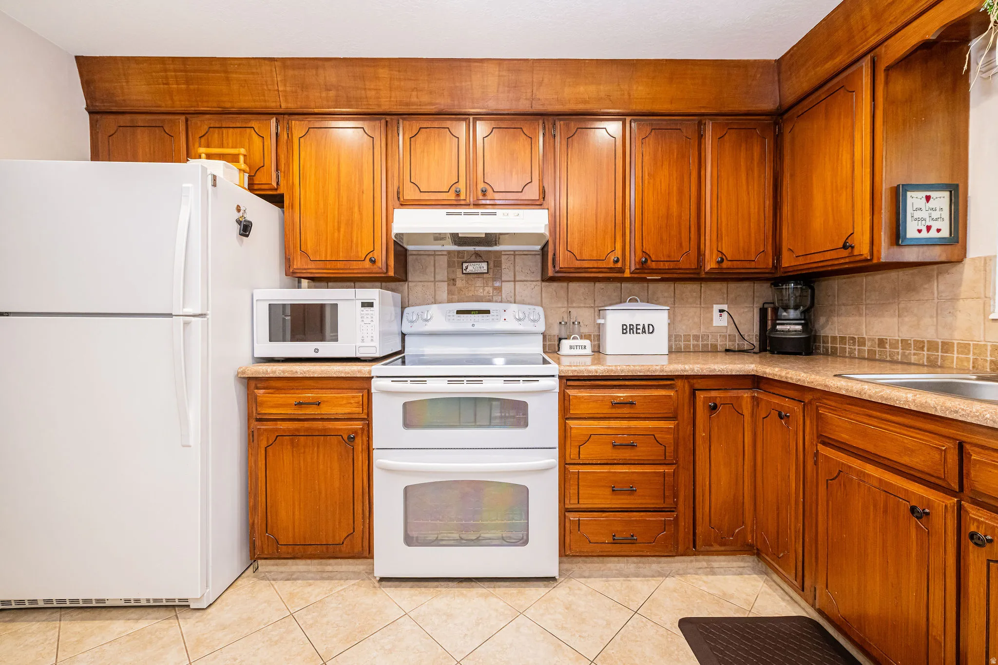 Kitchen featuring white appliances, brown cabinets, tasteful backsplash, under cabinet range hood, and light tile patterned floors