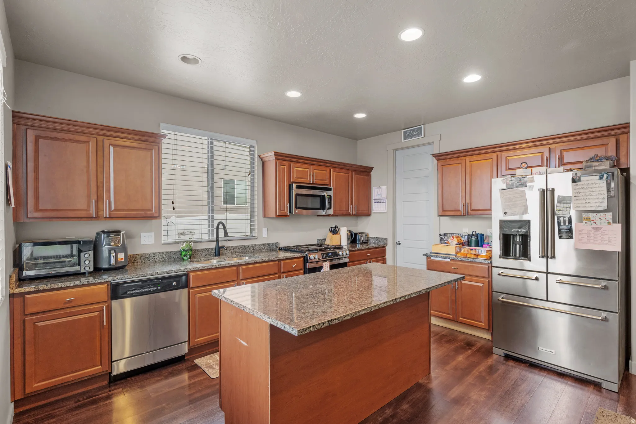 Kitchen with stainless steel appliances, wood finish cabinets, light stone countertops, a kitchen island, and dark wood finished floors