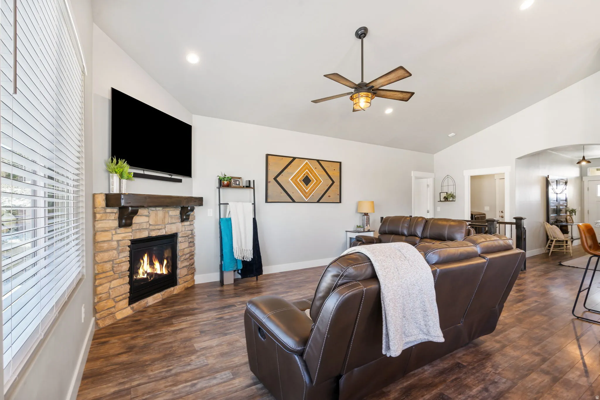 Living area with a ceiling fan, arched walkways, a stone fireplace, dark wood-style flooring, and lofted ceiling