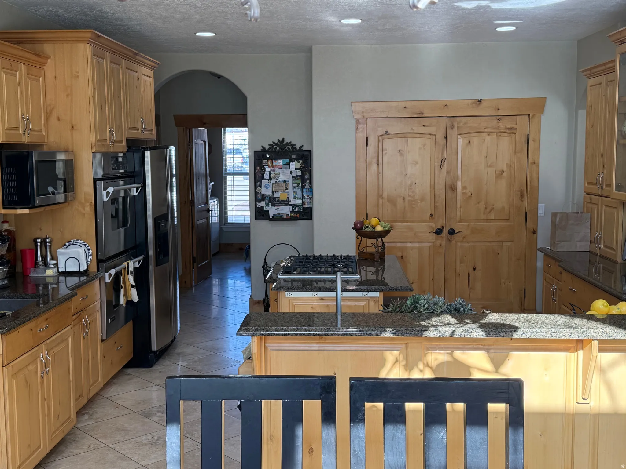 Kitchen featuring dark stone countertops, arched walkways, stainless steel appliances, light tile patterned flooring, and wood finish cabinetry