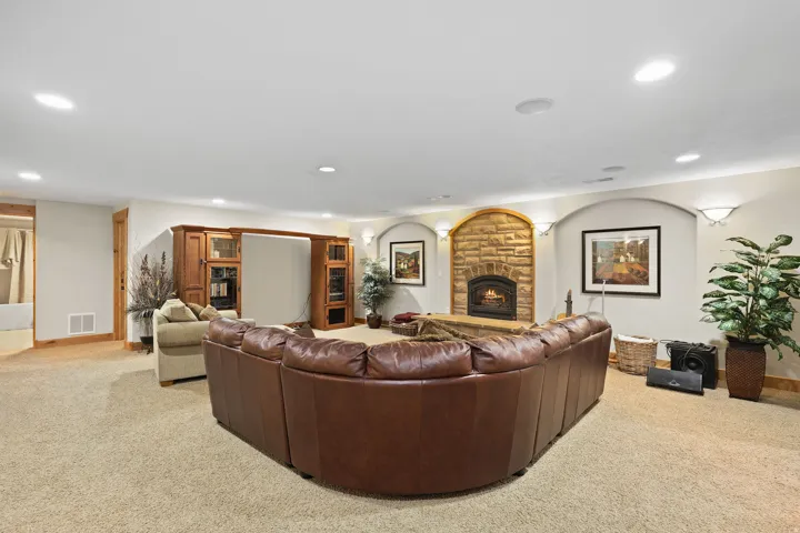 Spacious carpeted room featuring a stone fireplace with an arched surround, recessed ceiling lighting, wall sconces, and a built-in media center with glass-front cabinetry