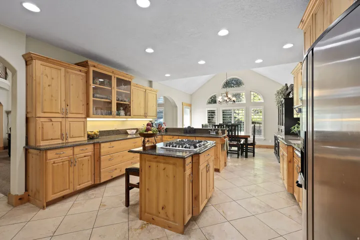 Spacious kitchen featuring warm wood cabinetry, dark stone countertops, stainless steel appliances, a center island with a gas cooktop, and large arched windows with plantation shutters