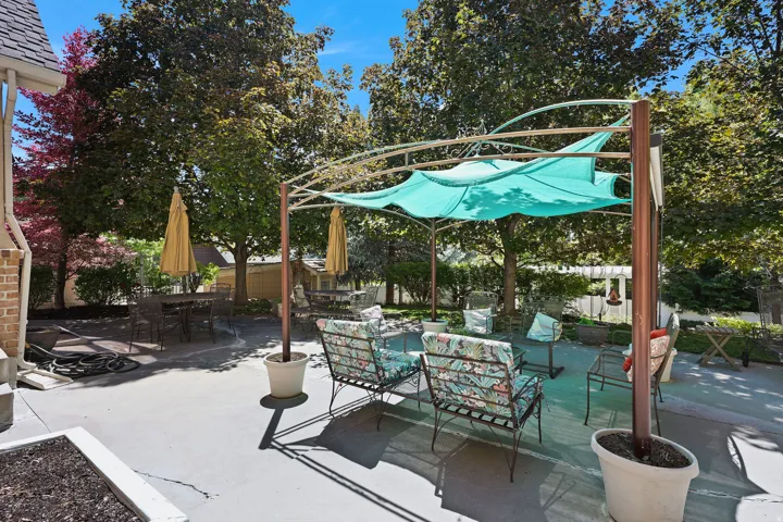 Spacious patio featuring a metal pergola with a fabric shade, extensive paved surfacing, and mature trees providing natural canopy