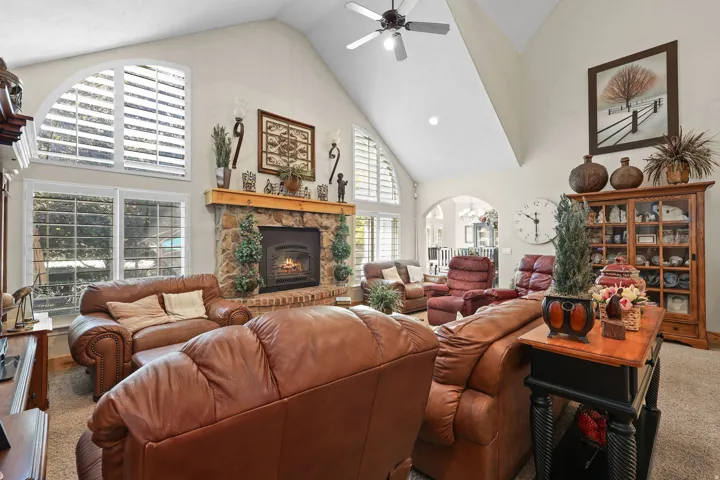 Vaulted ceiling living area featuring a stone fireplace with wood mantel, arched windows with shutters, ceiling fan, and carpeted flooring
