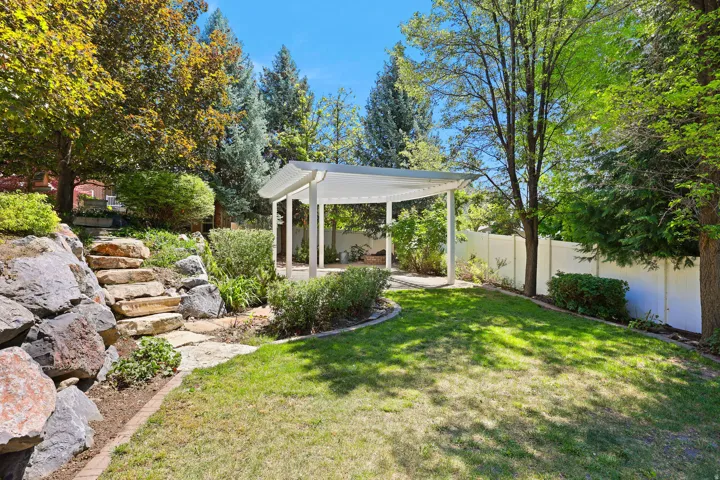 Expansive backyard featuring a white pergola structure, a large grassy area, a rock retaining wall with stone steps, mature trees, and a white privacy fence