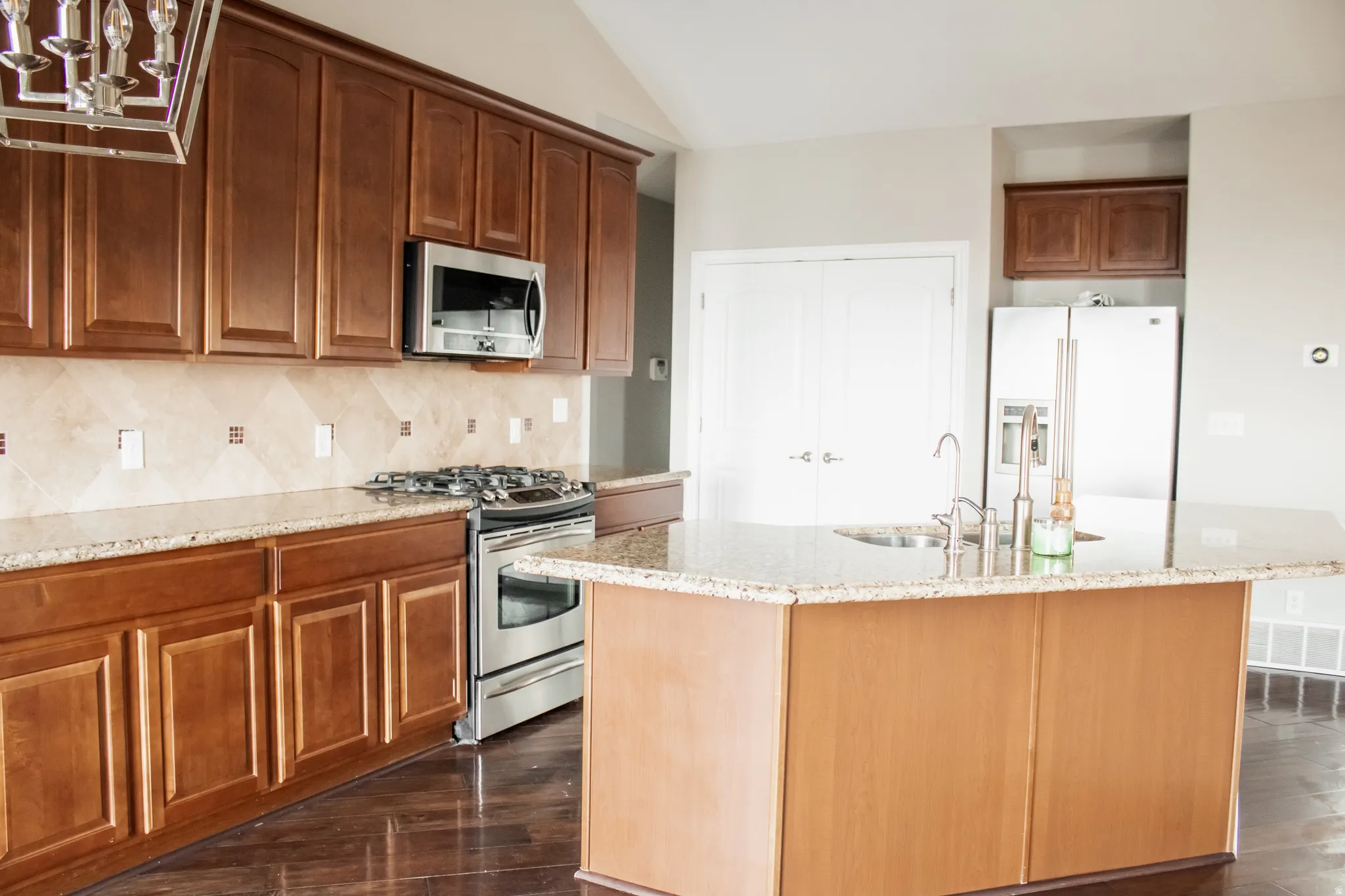 Kitchen featuring appliances with stainless steel finishes, light stone countertops, dark wood finished floors, vaulted ceiling, and an island with sink