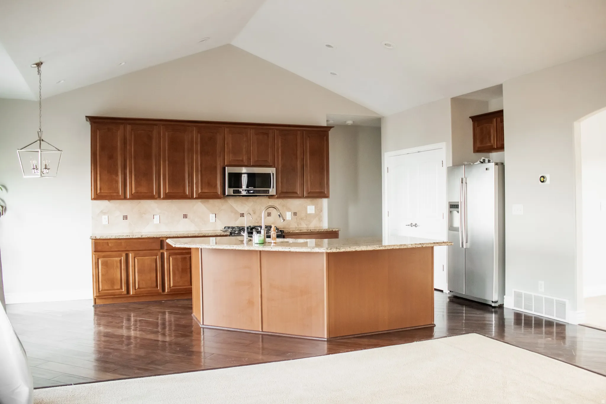 Kitchen with appliances with stainless steel finishes, light stone counters, a kitchen island with sink, dark wood finished floors, and lofted ceiling