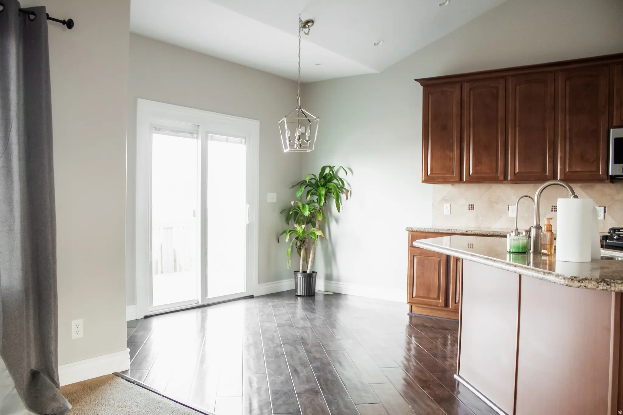 Kitchen with hanging light fixtures, vaulted ceiling, light stone countertops, dark wood-type flooring, and backsplash