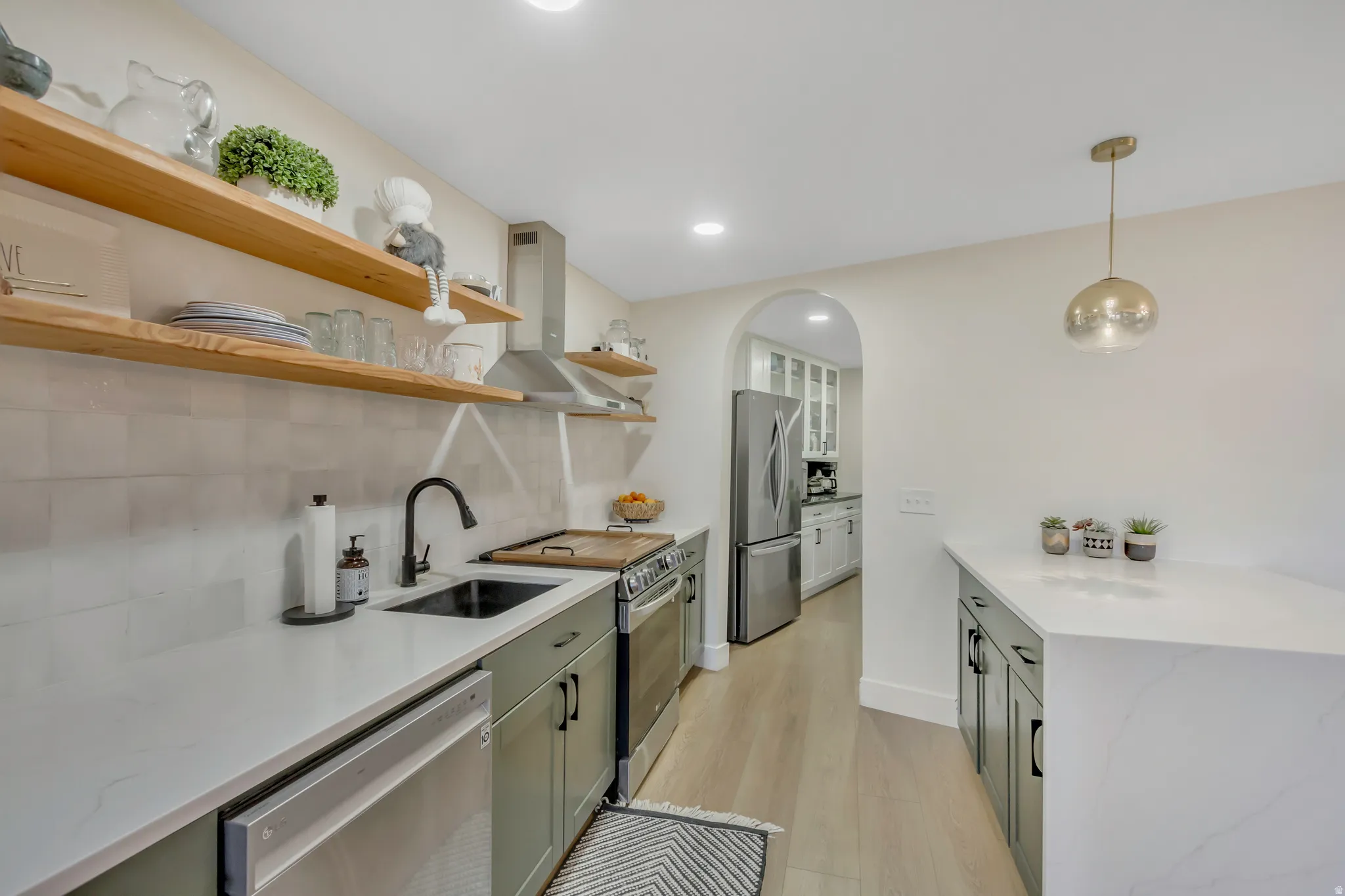 Kitchen with pendant lighting, appliances with stainless steel finishes, wall chimney range hood, light stone counters, and light wood-style flooring