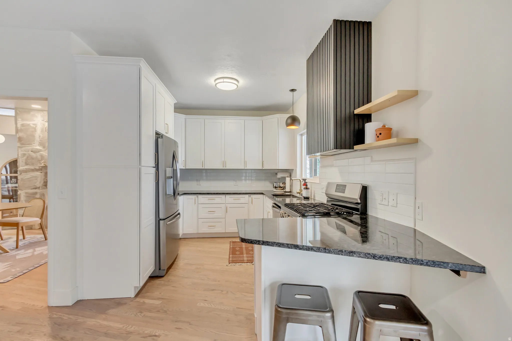 Kitchen with dark stone countertops, light wood finished floors, hanging light fixtures, open shelves, and a kitchen bar
