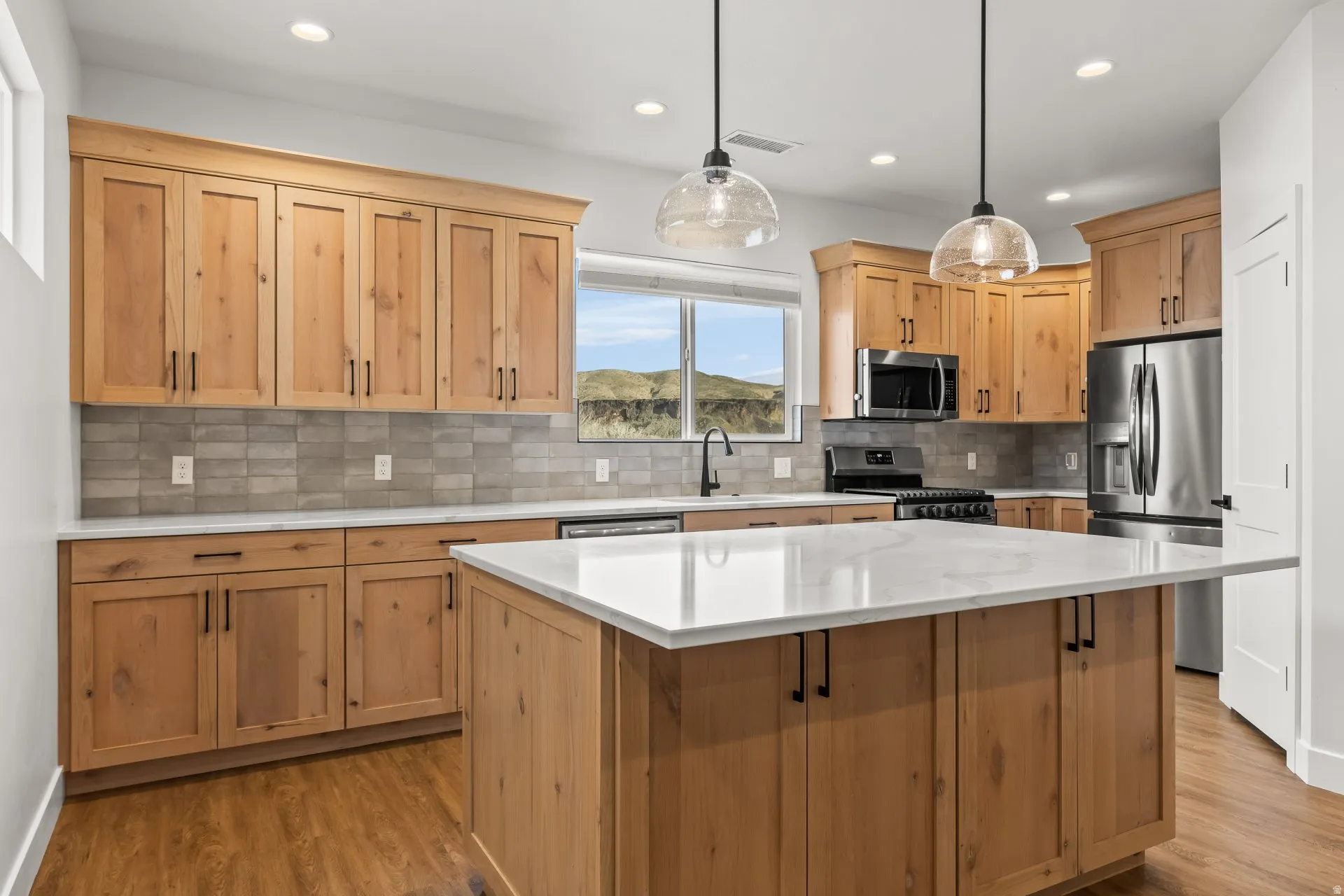 Kitchen with light wood finish cabinetry, stainless steel appliances, decorative light fixtures, light stone countertops, and a kitchen island
