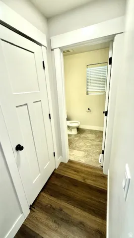 Hallway featuring wood-finish flooring, white panel doors with black hardware, and a doorway opening to a room with a window, white blinds, and light-toned flooring