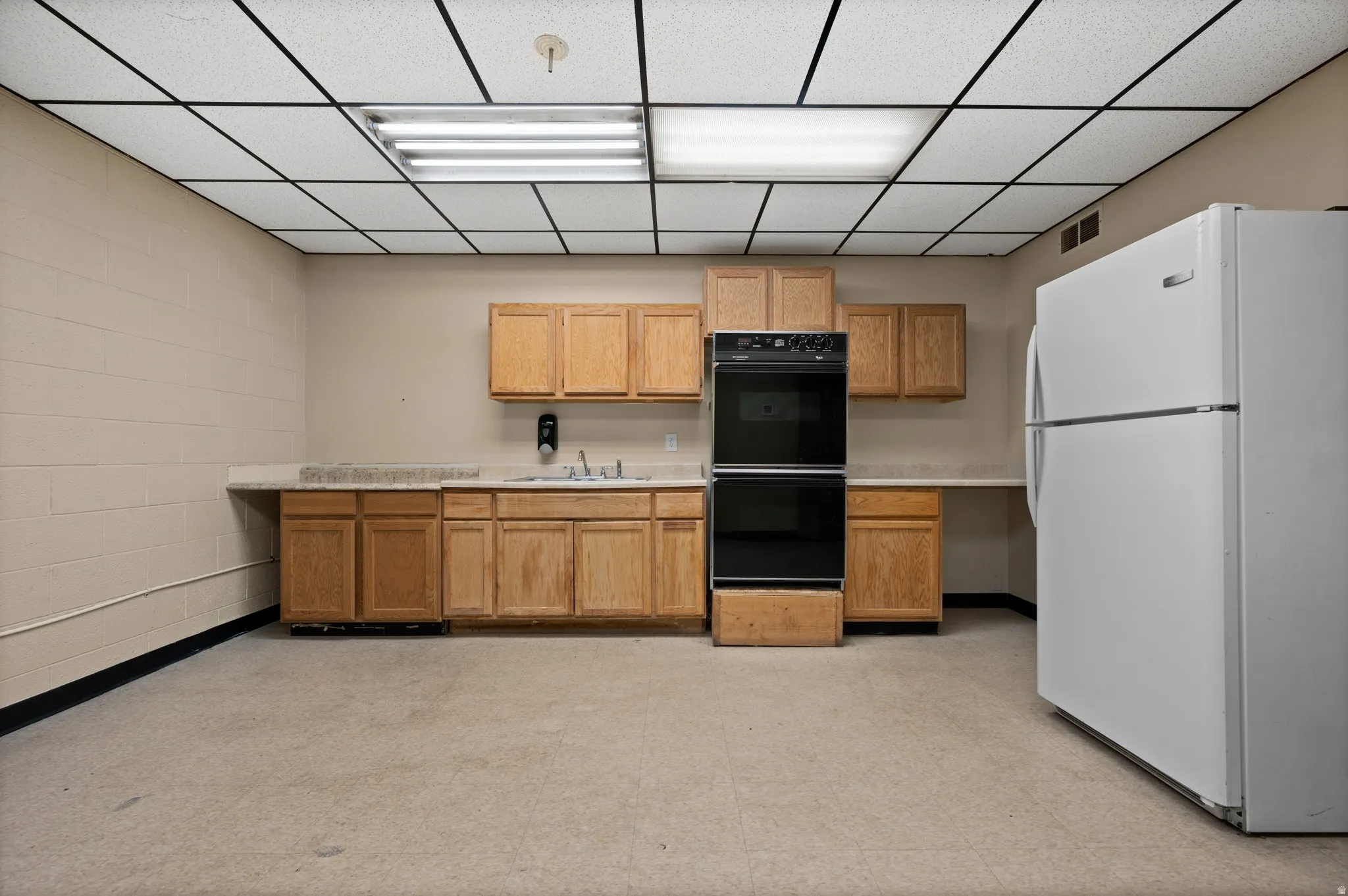 Kitchen with freestanding refrigerator, light countertops, a drop ceiling, and brown cabinets