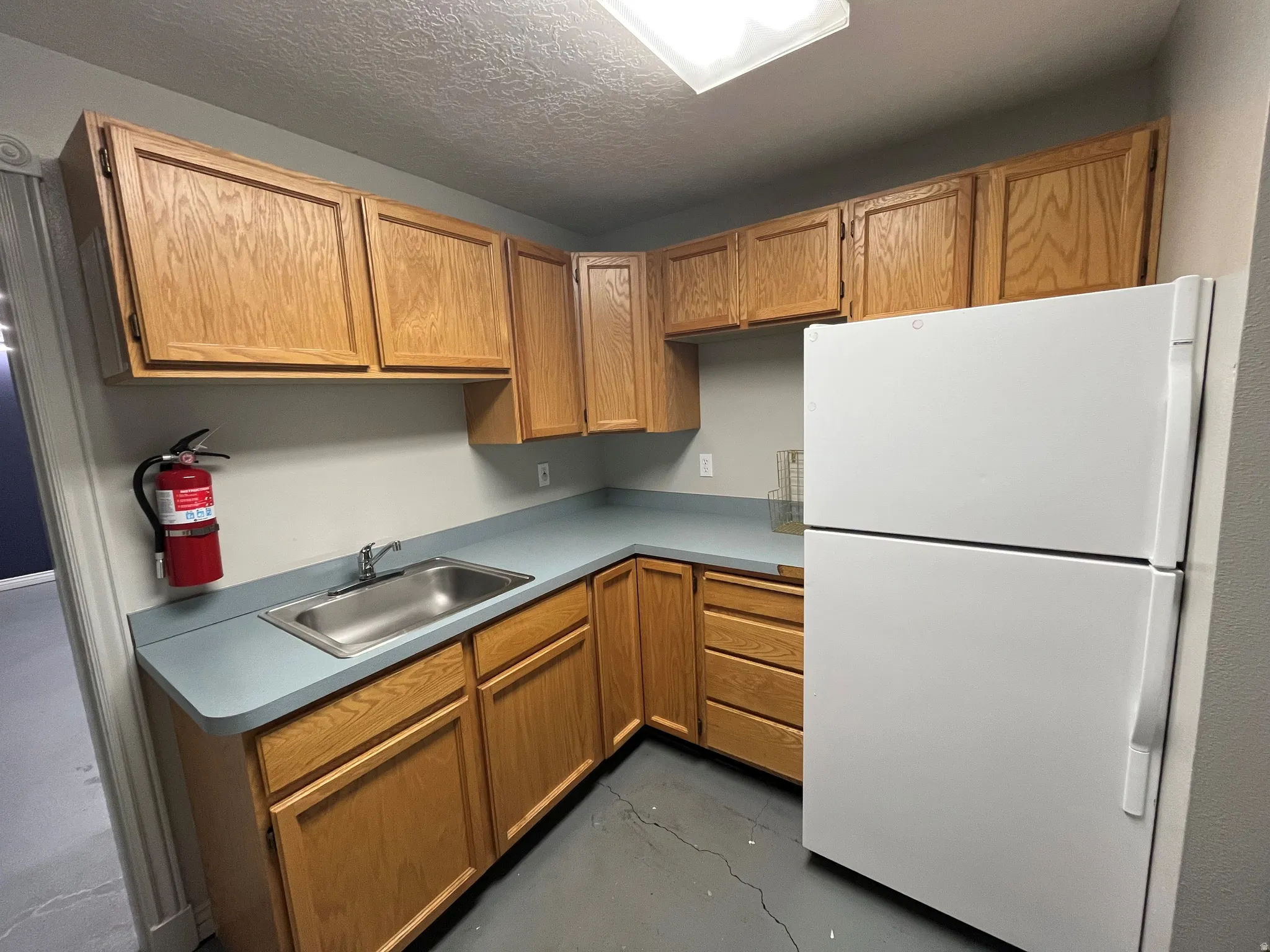 Kitchen with freestanding refrigerator, light countertops, a textured ceiling, wood finish cabinets, and finished concrete flooring