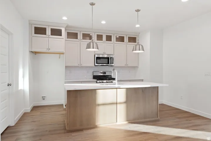 Kitchen with light wood-style flooring, stainless steel appliances, glass insert cabinets, an island with sink, and two tone cabinets