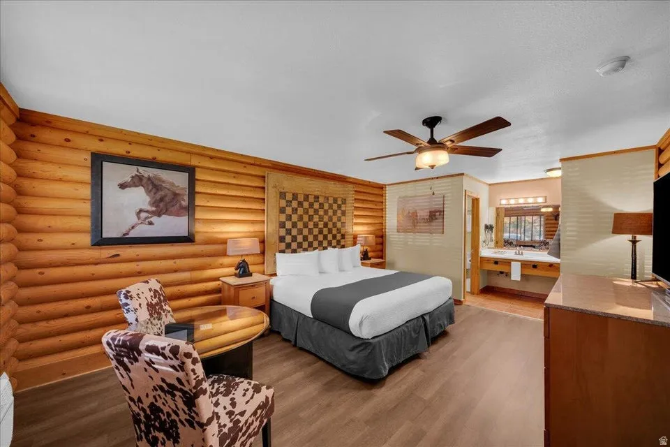 Bedroom with log walls, dark wood-type flooring, ensuite bath, and ceiling fan