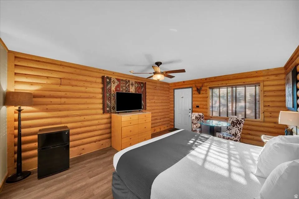 Bedroom with rustic walls, freestanding refrigerator, wood finished floors, and ceiling fan