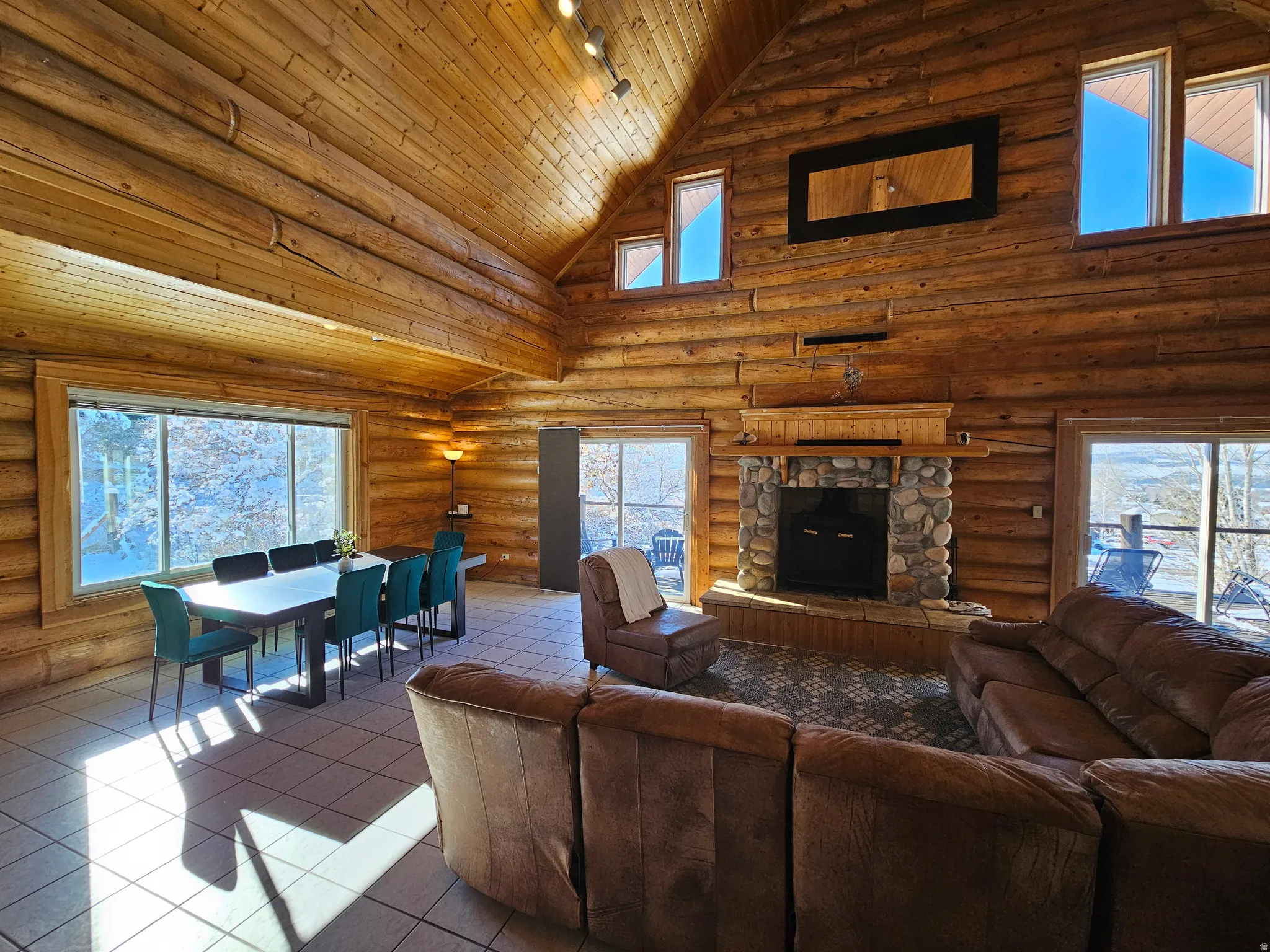 Tiled living area featuring healthy amount of natural light, a vaulted wood ceiling, log walls, rail lighting, and a stone fireplace