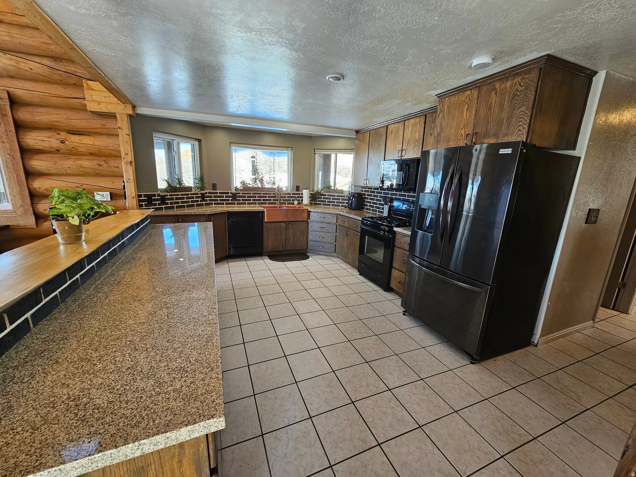 Kitchen featuring black appliances, rustic walls, dark wood finish cabinets, a textured ceiling, and light tile patterned flooring