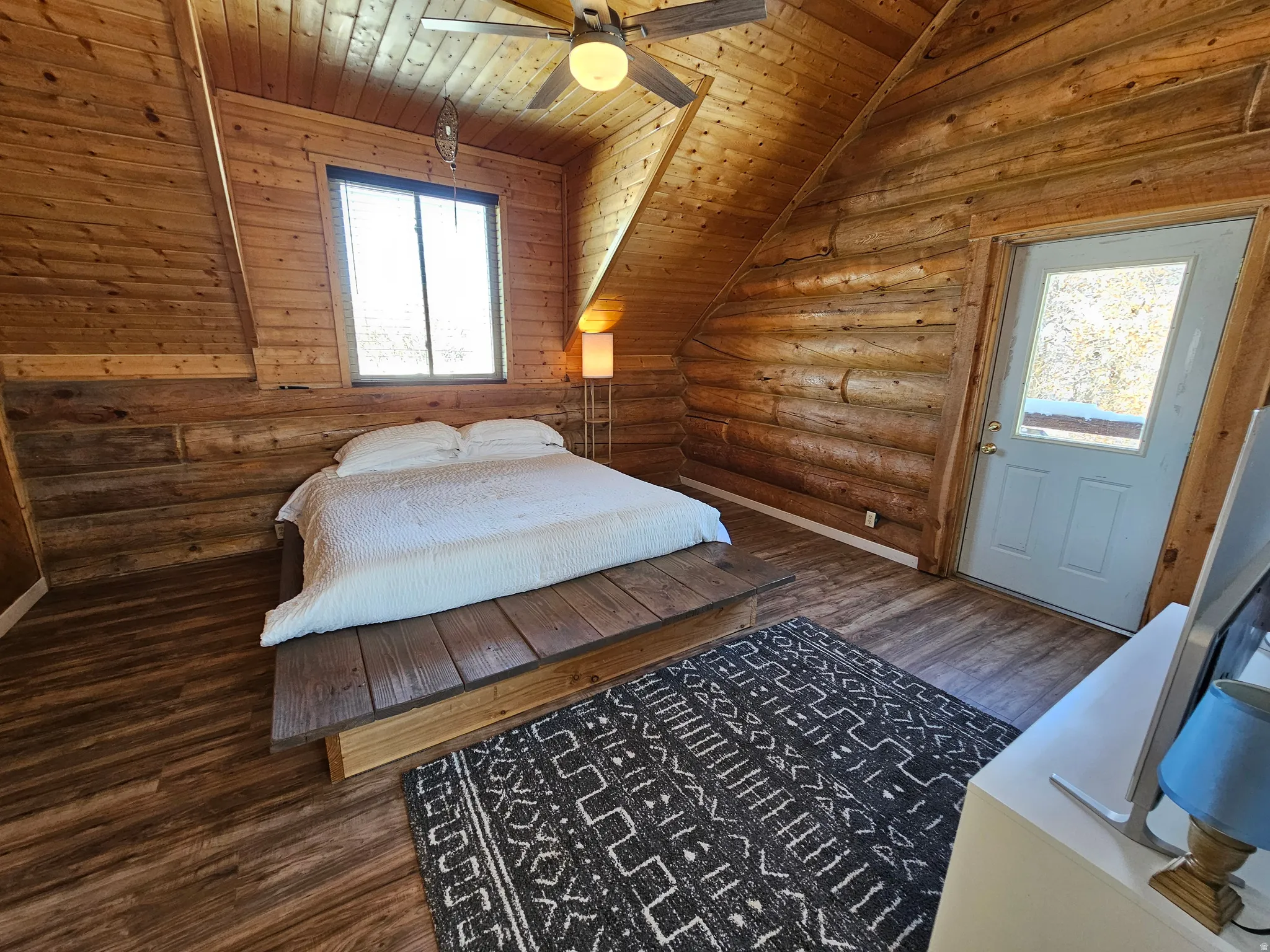 Bedroom with a vaulted wooden ceiling, rustic walls, dark wood-style floors, and a ceiling fan